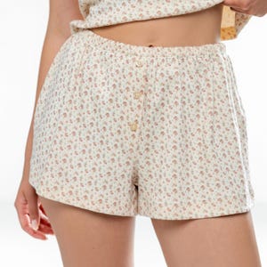 May include: Cream-colored shorts with a floral pattern. The shorts have a button-up front and an elastic waistband. The fabric is covered in small, pink and blue flowers. The shorts are part of a matching set.