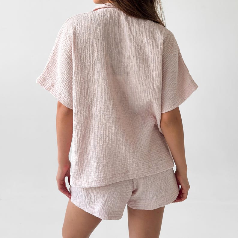 May include: A light pink, short-sleeved pajama set. The set includes a collared, button-down shirt and matching shorts. The fabric has a textured, crinkled appearance. The outfit is made of a soft, lightweight material.