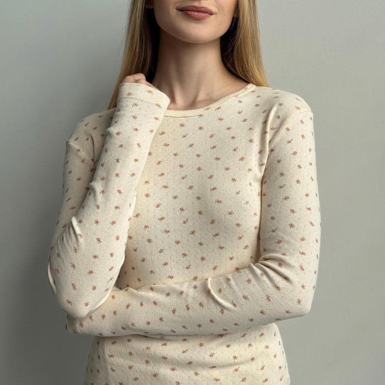 K&ouml;nnte beinhalten: Cremefarbenes Langarmshirt mit zartem Blumenmuster in einem ged&auml;mpften r&ouml;tlich-braunen Farbton. Das Shirt hat einen Rundhalsausschnitt und eine figurbetonte Silhouette, geeignet zum Unterziehen oder solo tragen. Der Stoff wirkt weich und leicht.