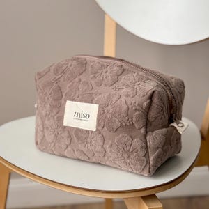 May include: A taupe-colored zippered pouch made of textured terry cloth fabric featuring an embossed floral pattern. A rectangular white fabric label is sewn onto the front, displaying the black text miso with smaller text underneath.