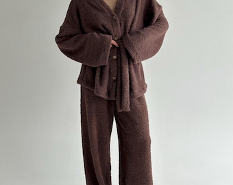 May include: A brown, fuzzy pajama set. The set includes a button-down cardigan with a V-neck and wide sleeves, and matching wide-leg pants. The fabric appears soft and comfortable, perfect for lounging. The color is a rich, warm brown.