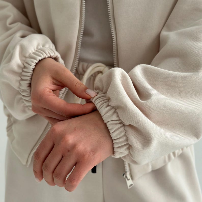 May include: A beige zip-up jacket with elastic cuffs and a matching drawstring waistband. The jacket is made of a soft, textured fabric. The image shows the jacket's details, including the zipper and the gathered sleeves.