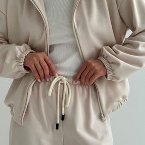 May include: A beige tracksuit set, featuring a zip-up jacket and matching drawstring pants. The jacket has a high collar and ribbed cuffs. The pants have a drawstring waist and a relaxed fit. The outfit is made of a soft, comfortable material.