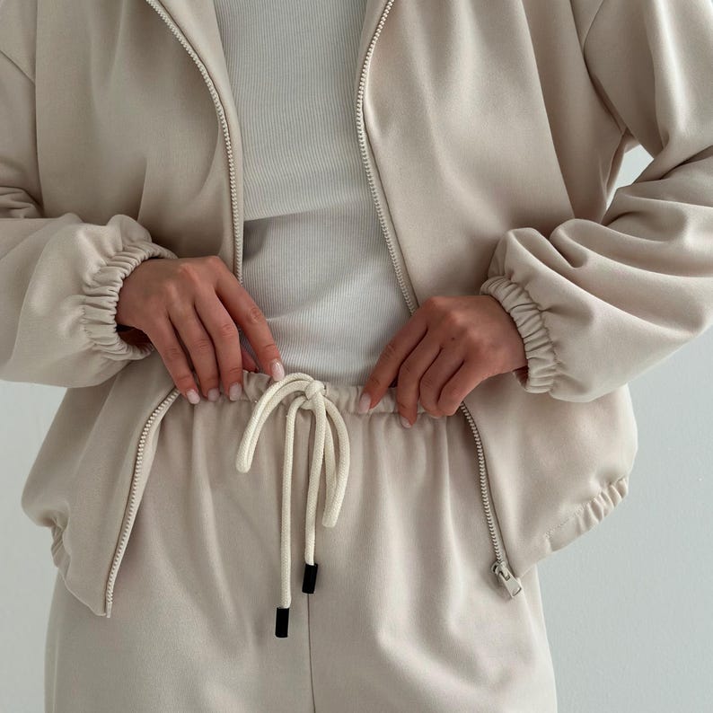 May include: A beige tracksuit set, featuring a zip-up jacket and matching drawstring pants. The jacket has a high collar and ribbed cuffs. The pants have a drawstring waist and a relaxed fit. The outfit is made of a soft, comfortable material.