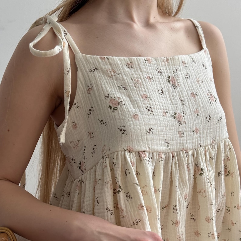 Op de afbeelding: Cr&egrave;mekleurige jurk met bloemenprint en strikbandjes. De jurk heeft een vierkante halslijn en een gerimpelde rok. De stof heeft een getextureerde, gekreukelde uitstraling en het bloemenpatroon bevat roze en donkergroene accenten.