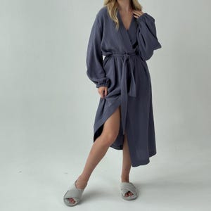 May include: A long, slate blue robe with a wrap-around design and a matching belt. The robe features long sleeves with gathered cuffs and a V-neckline. The fabric has a textured appearance. The model is wearing gray slippers.