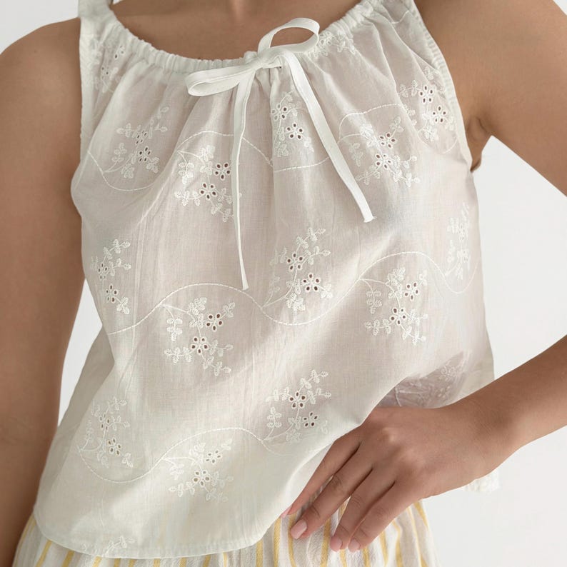 May include: A sleeveless white top featuring an eyelet floral pattern and a gathered neckline with a white ribbon tie. The fabric displays a repeating design of embroidered flowers and leaves arranged in horizontal wavy lines across the garment.