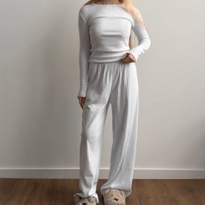 Puede incluir: Un conjunto de pijama blanco de punto acanalado. La parte superior de manga larga tiene cuello barco, y los pantalones son de pernera ancha con cintura elástica. La persona lleva zapatillas con forma de oso. El conjunto es de un material suave y cómodo.
