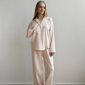 Op de afbeelding: Een lichtroze en wit gestreepte pyjamaset. De set bevat een overhemd met lange mouwen en knopen met een borstzak en een bijpassende broek met wijde pijpen. De stof heeft een getextureerde uitstraling.