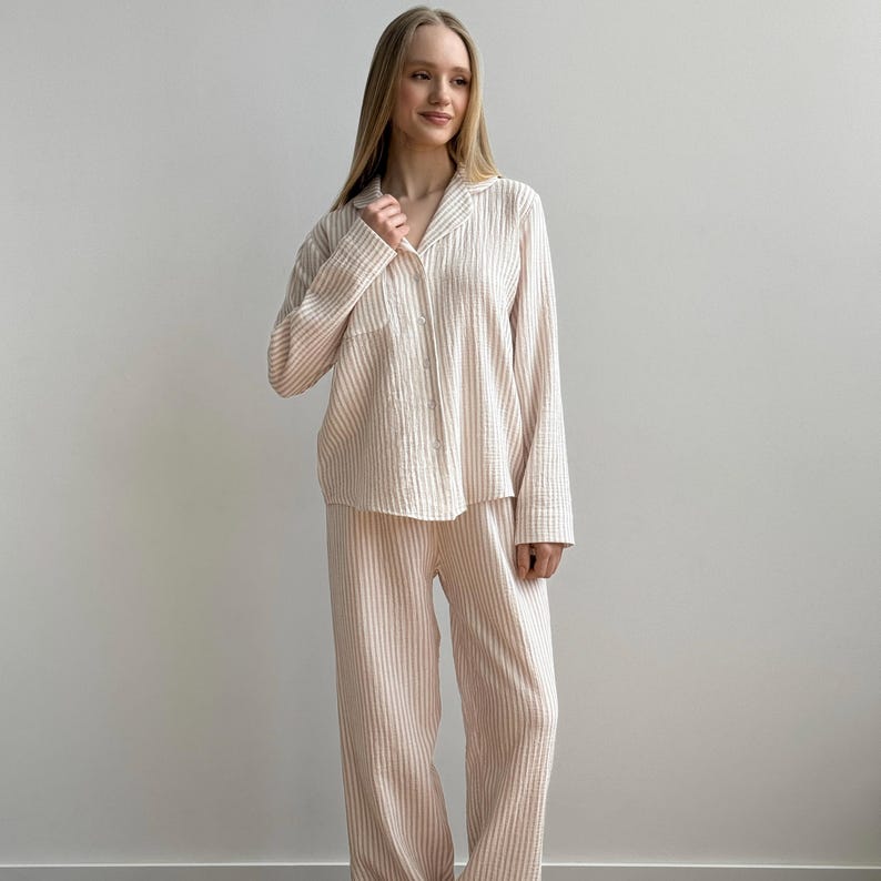 Op de afbeelding: Een lichtroze en wit gestreepte pyjamaset. De set bevat een overhemd met lange mouwen en knopen met een borstzak en een bijpassende broek met wijde pijpen. De stof heeft een getextureerde uitstraling.