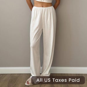 May include: Cream-colored, ribbed-knit pants with a matching top. The pants have a wide-leg design and an elastic waistband. The outfit is made of a soft, textured fabric. The text "ALL US TAXES PAID" is at the bottom of the image.