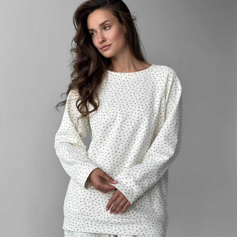 May include: Cream-colored long-sleeved sweatshirt with a small, repeating floral pattern. The sweatshirt has a crew neck and raglan sleeves. The fabric appears soft and comfortable, suitable for casual wear. The overall style is relaxed and cozy.