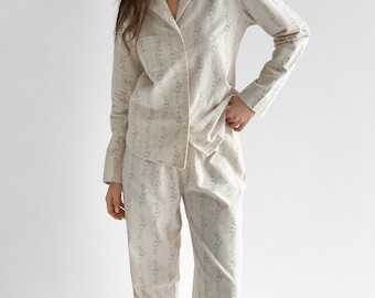 May include: A two-piece pajama set in a light cream color featuring a vertical pattern of small, muted blue floral vines. The long-sleeved button-down shirt has a notched collar and a single chest pocket, paired with matching long, straight-leg pants.