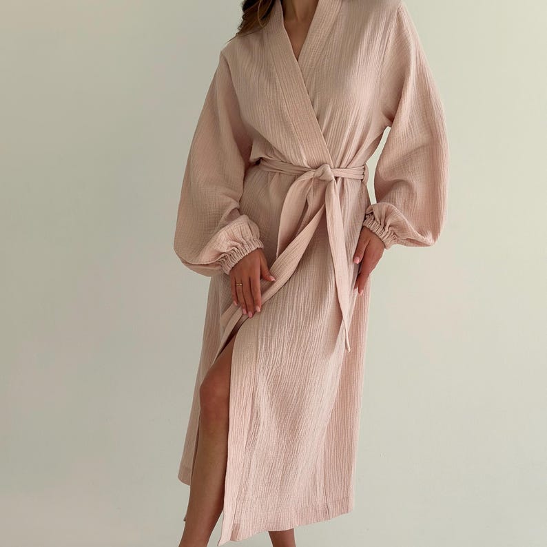May include: A light pink robe with long, voluminous sleeves and a matching tie belt. The robe has a wrap-around design and a high side slit. The fabric appears to be textured, and the robe is knee-length.