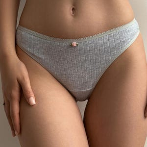 May include: Light gray ribbed underwear with a delicate floral accent. The underwear features a scalloped trim along the waistline and a small pink rose detail at the center front. The fabric has a textured appearance.