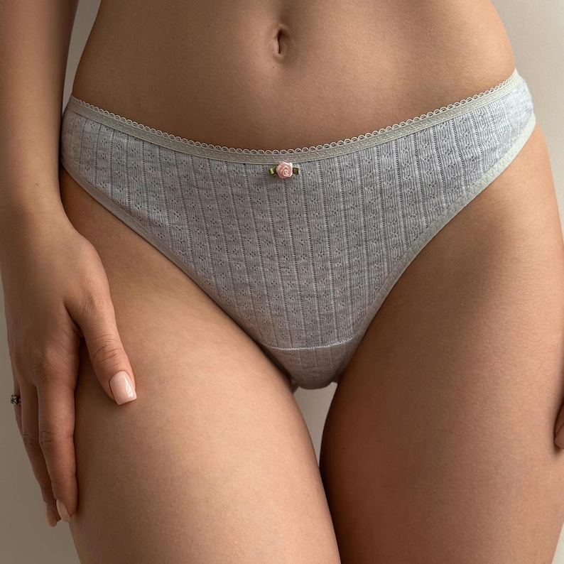 May include: Light gray ribbed underwear with a delicate floral accent. The underwear features a scalloped trim along the waistline and a small pink rose detail at the center front. The fabric has a textured appearance.