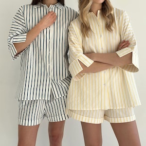 May include: Two sets of striped pajamas. One set is white with dark green stripes, the other is white with yellow stripes. Each set includes a button-down shirt with three-quarter sleeves and matching shorts. The pajamas are made of a lightweight fabric.