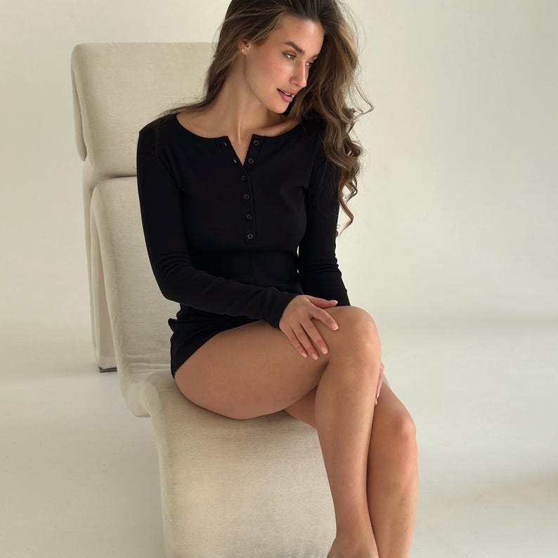 May include: Black long-sleeved henley top and matching shorts. The top features a scoop neck and a button-down placket. The outfit is made of a ribbed knit fabric. The model is sitting on a beige chair.