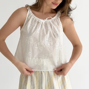 May include: A white sleeveless top featuring an eyelet lace floral pattern with wavy embroidered lines and a drawstring neckline tie. The garment is paired with yellow and white vertically striped bottoms, creating a light and airy appearance.