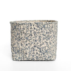 May include: A square, fabric storage basket with a floral pattern in blue and white. The basket is quilted with vertical stitching and has a neutral-colored interior. The basket is designed for organizing and storage.