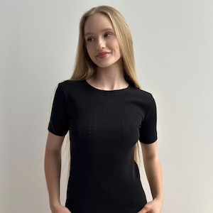 May include: A black, short-sleeved top with a ribbed texture. The top has a crew neck and a fitted shape. The fabric appears to be a soft knit material. The model is wearing the top.