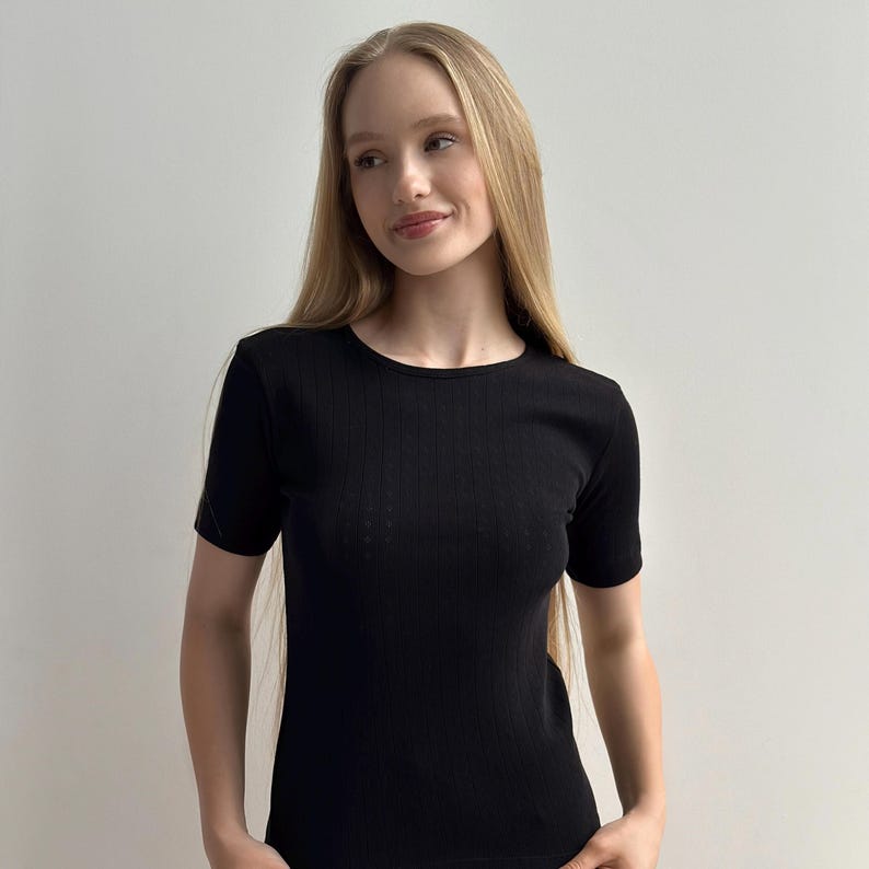 May include: A black, short-sleeved top with a ribbed texture. The top has a crew neck and a fitted shape. The fabric appears to be a soft knit material. The model is wearing the top.