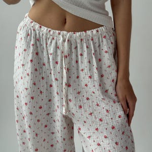 Puede incluir: Pantalones de pijama blancos con un estampado floral rojo. Los pantalones tienen cintura con cordón y un ajuste holgado y cómodo. La tela parece ser una mezcla ligera de algodón o lino.