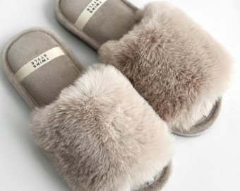 May include: A pair of plush, light brown slippers with a faux fur upper. The slippers have a soft, padded insole and a label that reads "TWINS STYLE". The open-toe design and fluffy texture suggest comfort and relaxation.