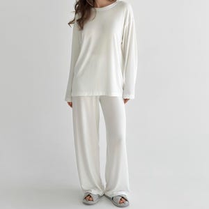 May include: A solid off-white long-sleeve shirt and matching wide-leg trousers set. The shirt features a crew neckline and long sleeves, while the trousers have a relaxed, loose fit that falls straight to the floor, creating a cohesive monochromatic look.