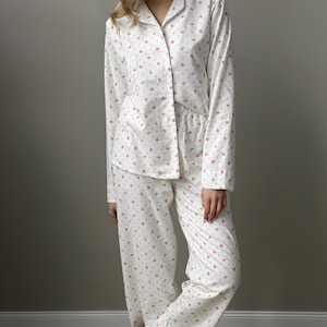 May include: White pajama set with a delicate floral pattern in red. The set includes a button-down shirt with a collar and long sleeves, and matching pants. The fabric appears soft and comfortable, suitable for sleepwear.