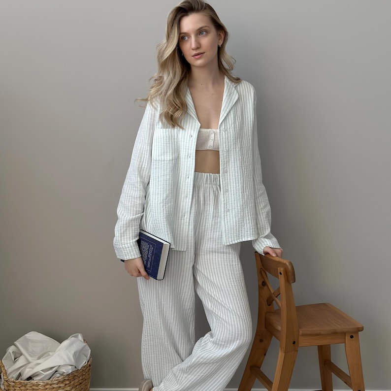 Op de afbeelding: Een lichtblauw en wit gestreepte pyjamaset, bestaande uit een overhemd met knopen, een broek en een bralette. De set is gemaakt van een lichte stof. Een persoon houdt een blauw boek vast en leunt tegen een houten stoel.