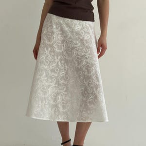 May include: A white midi skirt with an intricate, all-over lace pattern. The skirt is paired with a brown top. The skirt falls to mid-calf and has a slight A-line shape. The lace design adds a delicate and feminine touch to the garment.