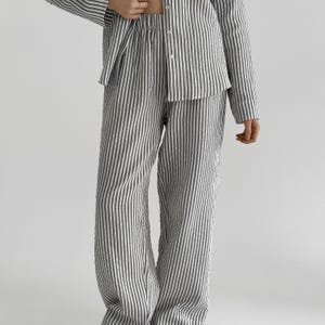 Op de afbeelding: Een tweedelige pyjamaset met een verticaal streepjespatroon. De set bestaat uit een overhemd met knopen en een bijpassende broek. De strepen zijn donkergrijs of zwart op een lichte achtergrond. De stof lijkt linnen of een vergelijkbaar lichtgewicht materiaal.