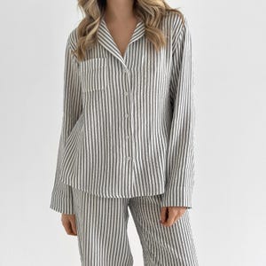 Op de afbeelding: Een gestreepte pyjamaset in grijstinten en wit. De lange mouwen blouse heeft een borstzak en knopen aan de voorkant. De bijpassende broek heeft een relaxte pasvorm. De stof lijkt licht van gewicht.