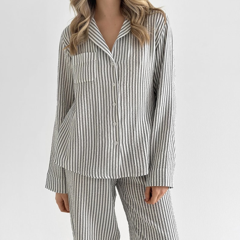 Op de afbeelding: Een gestreepte pyjamaset in grijstinten en wit. De lange mouwen blouse heeft een borstzak en knopen aan de voorkant. De bijpassende broek heeft een relaxte pasvorm. De stof lijkt licht van gewicht.