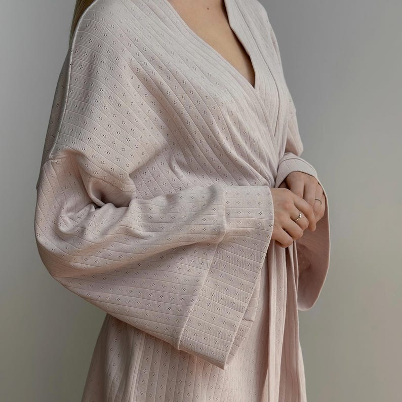 May include: A light pink, long-sleeved robe with a wrap-around design. The robe features a textured, patterned fabric and wide sleeves. The garment is tied at the waist with a matching belt. The robe is made of a soft, comfortable material.
