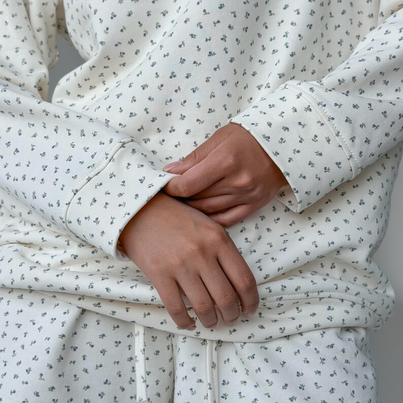 May include: Cream-colored sweatshirt and matching pants set with a delicate floral pattern in a muted blue-gray. The long-sleeved top has ribbed cuffs, and the pants have a drawstring. The outfit is made of a soft, comfortable material.