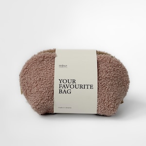 Könnte beinhalten: Eine ovale Tasche aus weichem, rosafarbenem Material. Die Tasche hat eine strukturierte, flauschige Außenseite und einen goldfarbenen Reißverschluss. Ein weißes Etikett um die Tasche zeigt "YOUR FAVOURITE BAG" in fetter schwarzer Schrift, mit der Marke "miso" darüber.