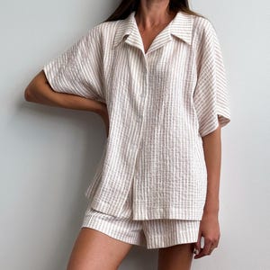 Peut inclure: Un ensemble de pyjama à rayures beiges et blanches. L'ensemble comprend une chemise boutonnée à manches courtes et un short assorti. Le tissu a un aspect texturé. La chemise a un col et le short a une taille élastique.