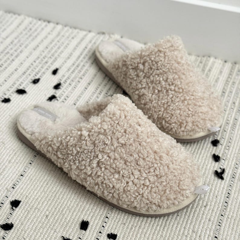 Peut inclure: Une paire de pantoufles beiges et duveteuses avec un design textur&eacute; et boucl&eacute;. Les pantoufles ont un aspect doux et moelleux et une couleur neutre, sugg&eacute;rant confort et chaleur. Elles sont pos&eacute;es sur une surface blanche cass&eacute; &agrave; motifs.