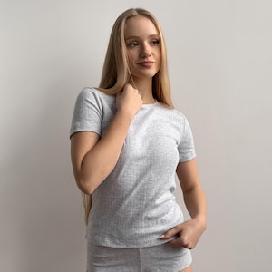 May include: A light grey ribbed knit t-shirt and matching shorts. The short-sleeved top has a crew neck. The fabric has vertical ribbing. The shorts are visible at the bottom of the image.