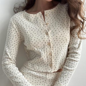 Pode incluir: Conjunto de cardigan e calças a condizer em creme. O cardigan de manga comprida tem uma frente abotoada e um decote redondo. O conjunto é coberto por um pequeno padrão floral repetido em cinza-esverdeado.