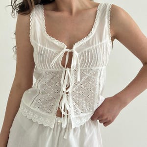 Puede incluir: Una blusa blanca sin mangas con escote redondo y ribete de encaje, pliegues verticales en la parte superior y una sección inferior con bordado floral de ojales. La parte delantera se abre con tres cintas finas y el dobladillo es festoneado.