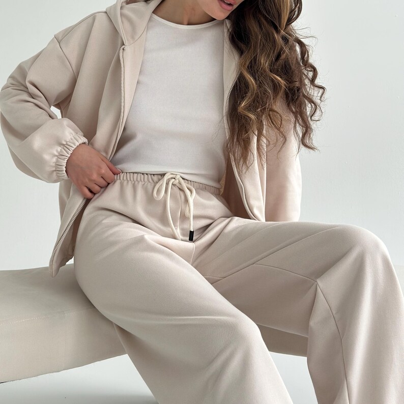 May include: A beige tracksuit set, featuring a zip-up hoodie, a white ribbed top, and matching drawstring pants. The hoodie has a hood and elastic cuffs. The pants have a drawstring waist. The outfit is made of a soft, comfortable material.