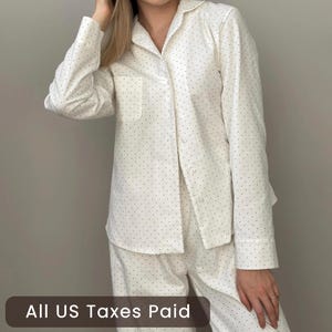 Op de afbeelding: Een pyjamashirt met lange mouwen en knoopsluiting in het wit met een klein zwart stippenpatroon. Het shirt heeft een klassieke kraag, een enkele borstzak aan de linkerkant en witte knopen die langs de voorzijde naar beneden lopen.