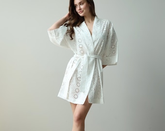 May include: A white short robe featuring an all-over eyelet lace pattern with circular floral cutouts. The garment has wide three-quarter length sleeves, a V-neckline, and a matching fabric tie belt that secures at the waist for a wrap-style closure.