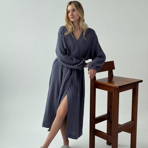 May include: A long, slate blue robe with a wrap-around design and a matching belt. The robe has long sleeves and a V-neck. The model is leaning against a wooden stool. The robe is made of a soft, flowing fabric.