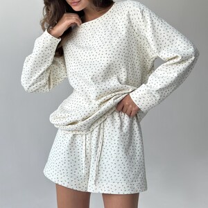 May include: Cream-colored loungewear set with a delicate floral pattern. The set includes a long-sleeved sweatshirt and matching shorts with a drawstring waist. The fabric appears soft and comfortable, perfect for relaxing at home.