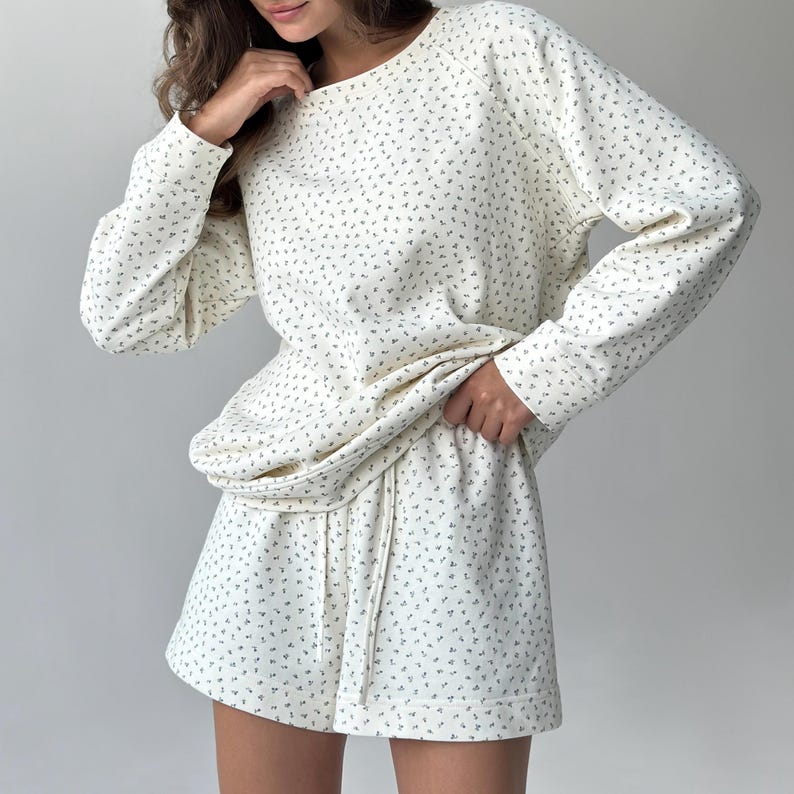 May include: Cream-colored loungewear set with a delicate floral pattern. The set includes a long-sleeved sweatshirt and matching shorts with a drawstring waist. The fabric appears soft and comfortable, perfect for relaxing at home.