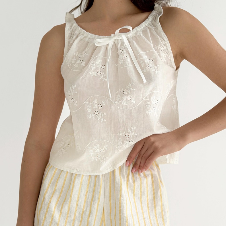 May include: A white sleeveless top featuring an eyelet floral pattern, a gathered neckline with a white ribbon tie, and a pair of yellow and white vertically striped shorts. The top has a scalloped hemline and a lightweight, semi-sheer appearance.